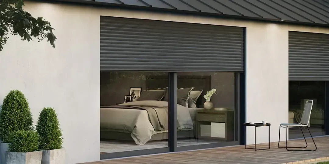 Vista externa de um quarto com persiana fechada. Prédio branco com detalhes em preto. Pequeno espaço para sentar ao ar livre.