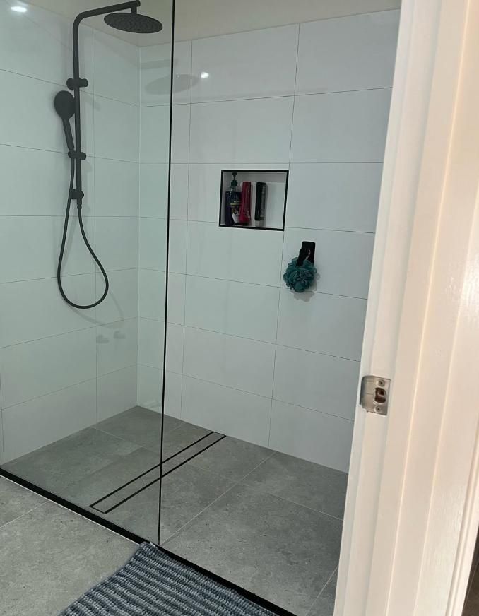Modern Shower With Black Fixtures, White Tiled Walls, And Gray Floor. Includes A Glass Door And A Recessed Shelf With Toiletries — Canberra Bathroom Solutions in Ngunnawal, ACT