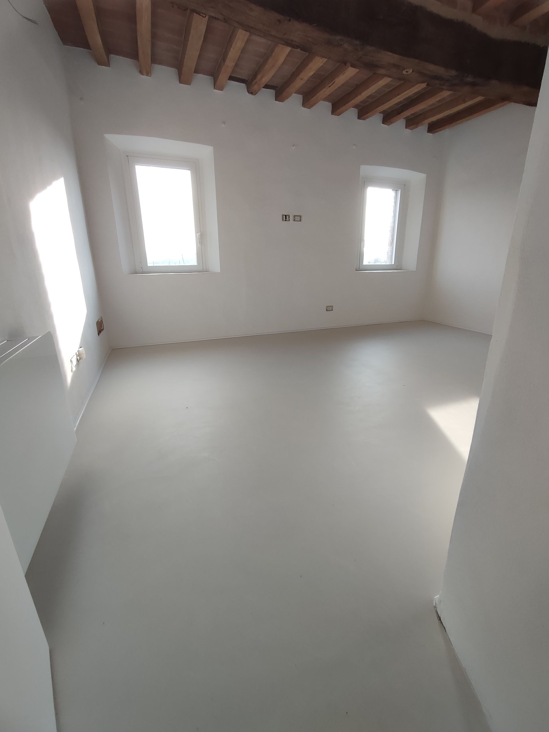 Stanza bianca e vuota, illuminata dalle finestre. Travi in legno al soffitto, pavimento chiaro e pareti nude.