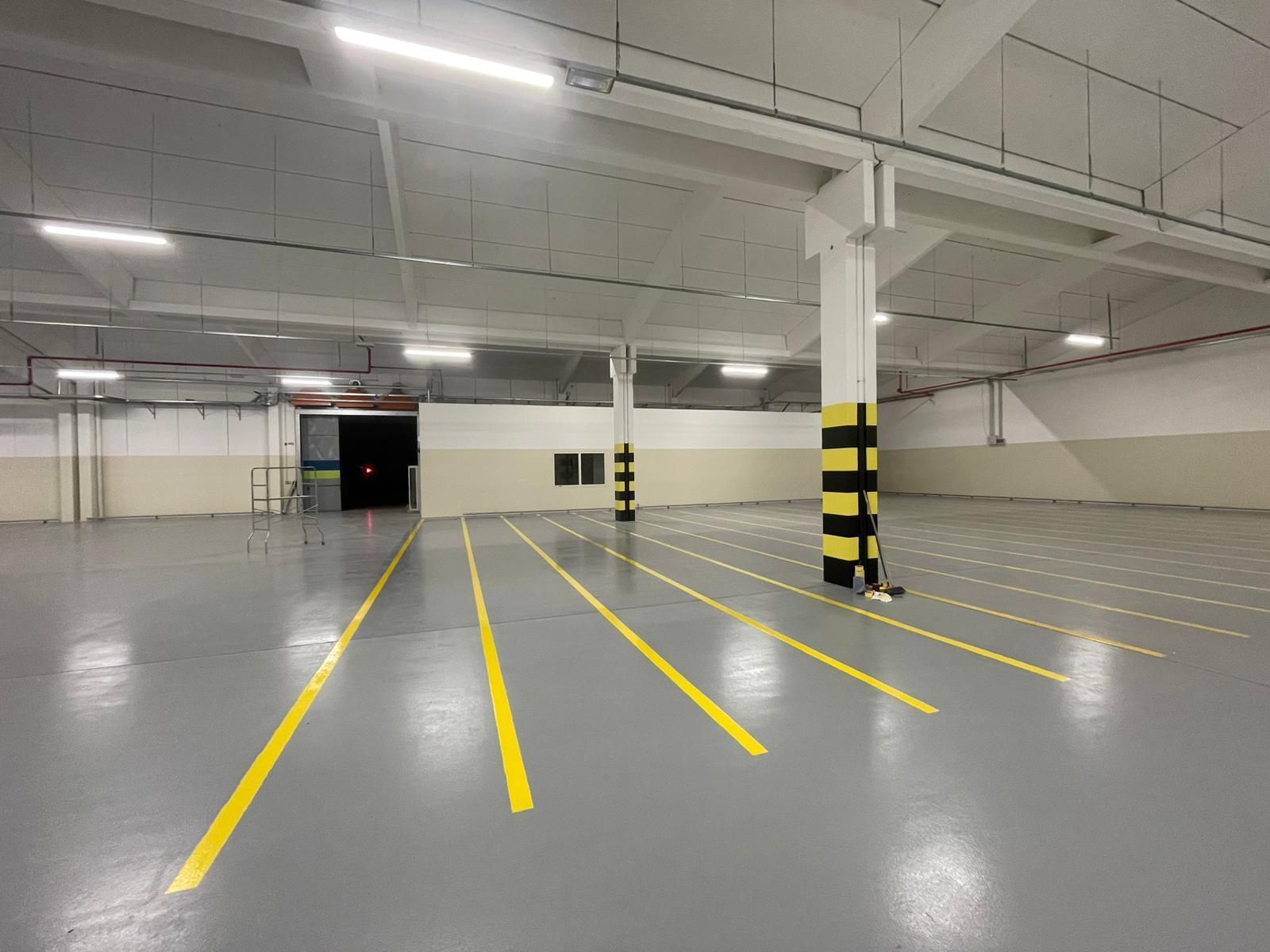 Spazio industriale vuoto con pavimento grigio, linee gialle, pilastri bianchi e luci sospese.