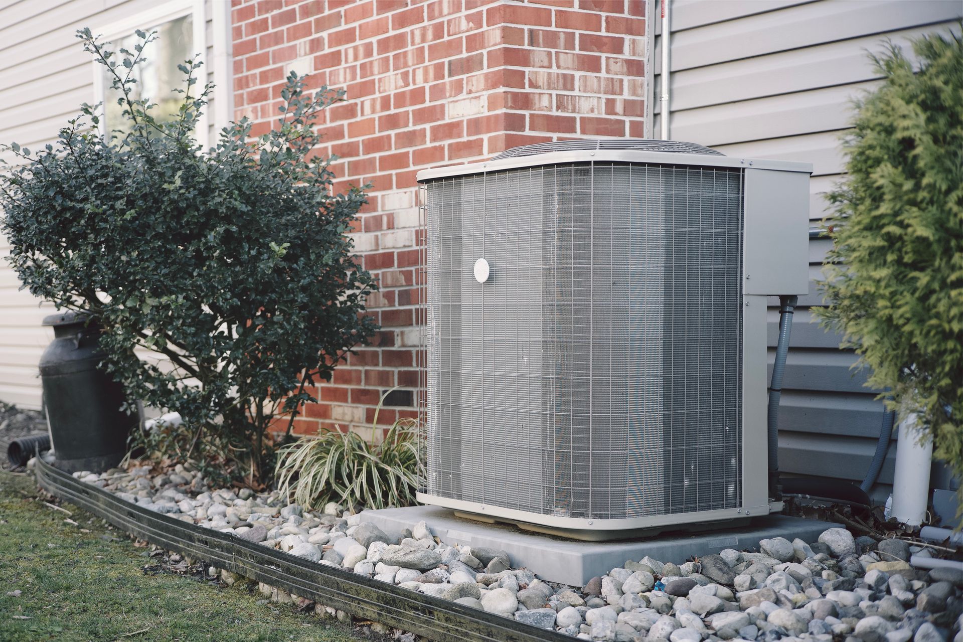 An outdoor air conditioning unit sits on a concrete pad next to a brick house, surrounded by gravel and green bushes.