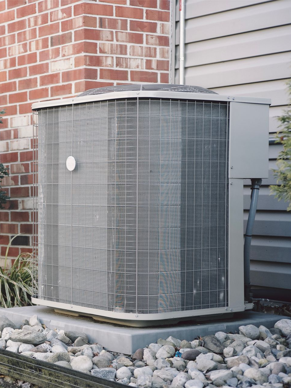 A gray residential air conditioning condenser unit sits on a concrete pad surrounded by landscaping rocks near a brick wall.