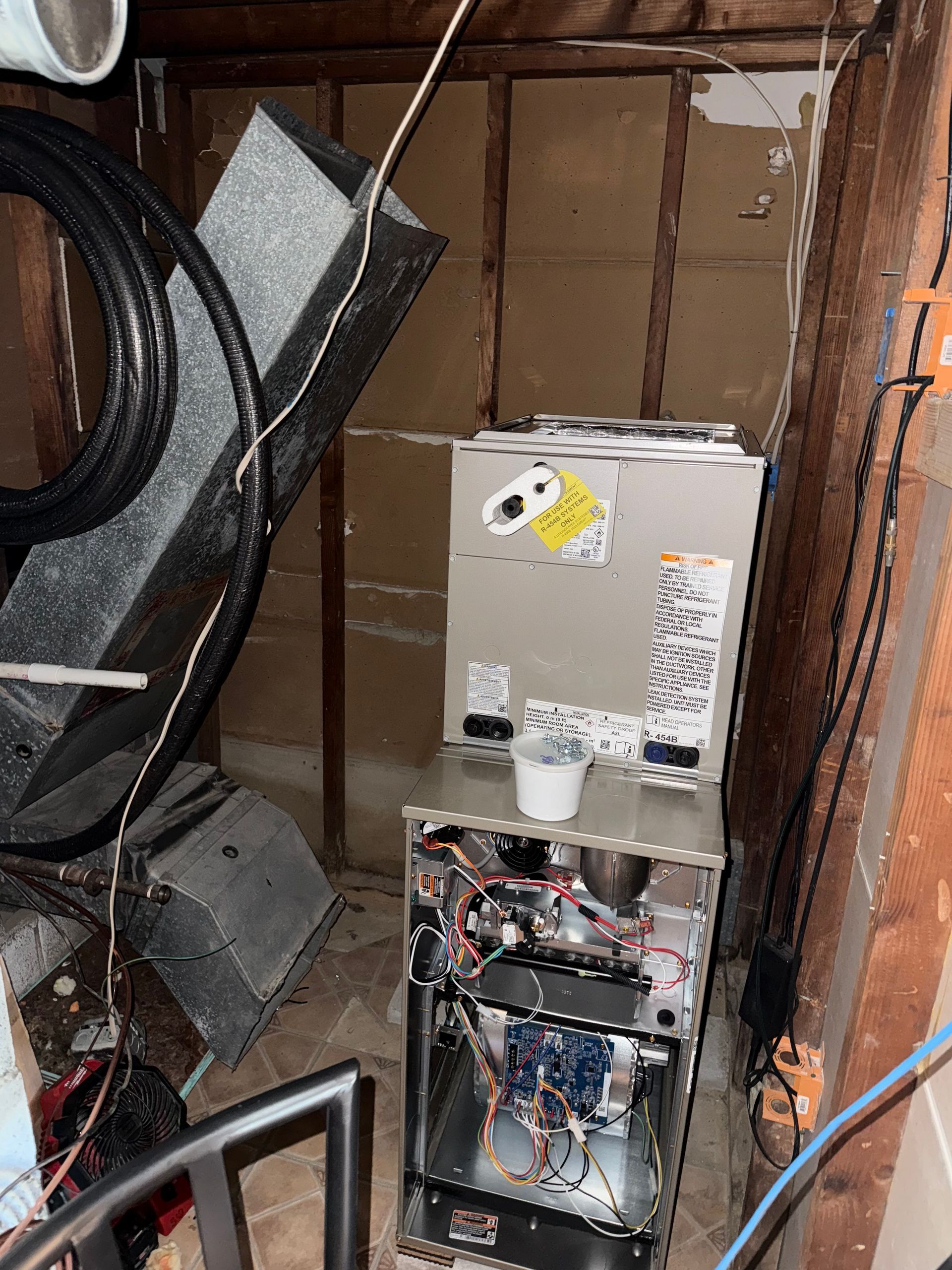 Furnace with open panels in a cluttered, unfinished attic. Wires and ductwork visible.