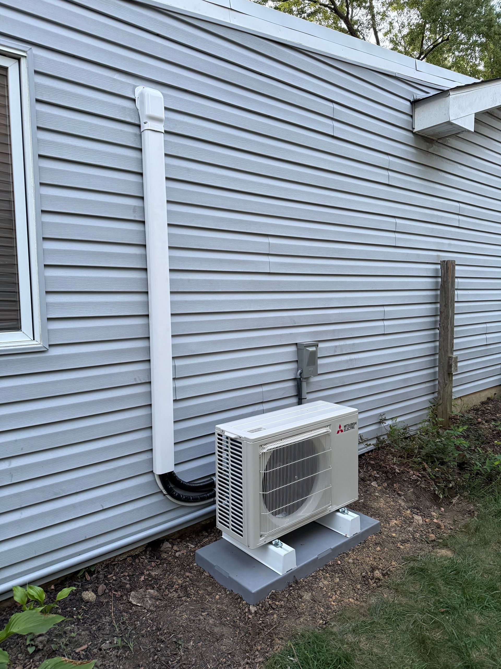 Outdoor air conditioning unit attached to a light blue siding building, with white vent pipe.