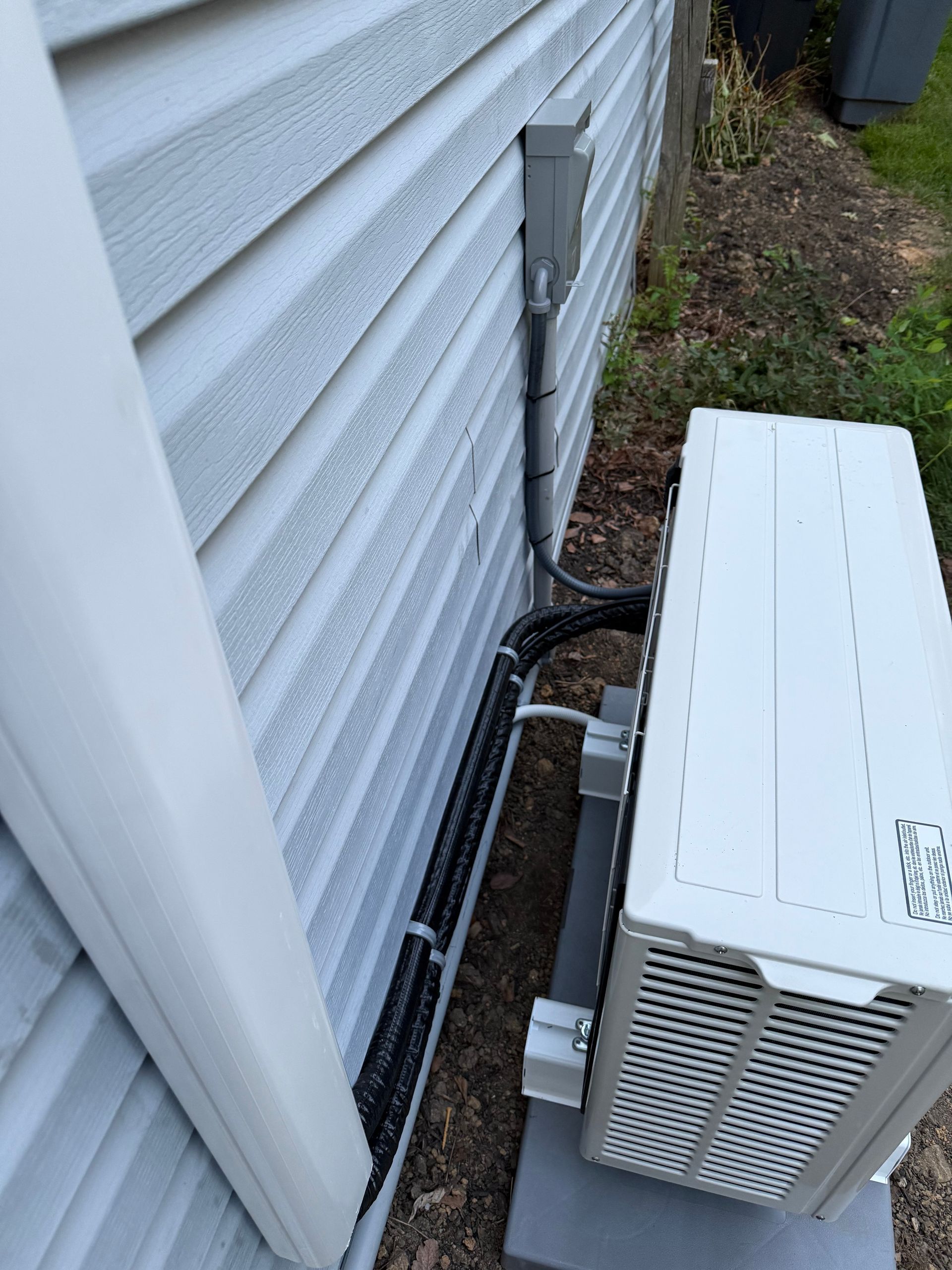 Air conditioner unit outside house, gray siding, electrical conduit along wall.