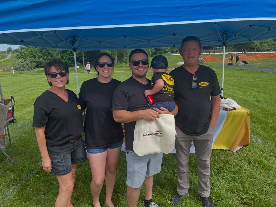 Five people standing under a blue canopy at an outdoor event. One man holds a baby. All wearing black shirts.