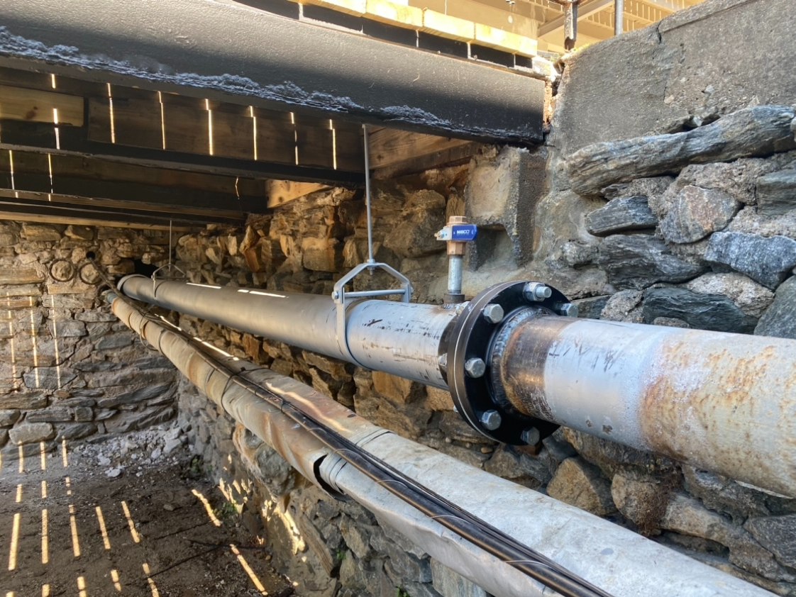 A metal pipe supported by a hanger and fitted with a flanged joint and valve runs alongside a rough stone wall.