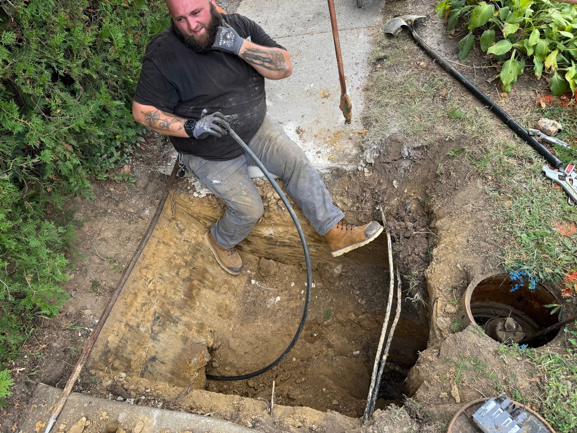 Man pulling cable from a deep outdoor hole. He wears work gloves, boots, and is surrounded by dirt and concrete.