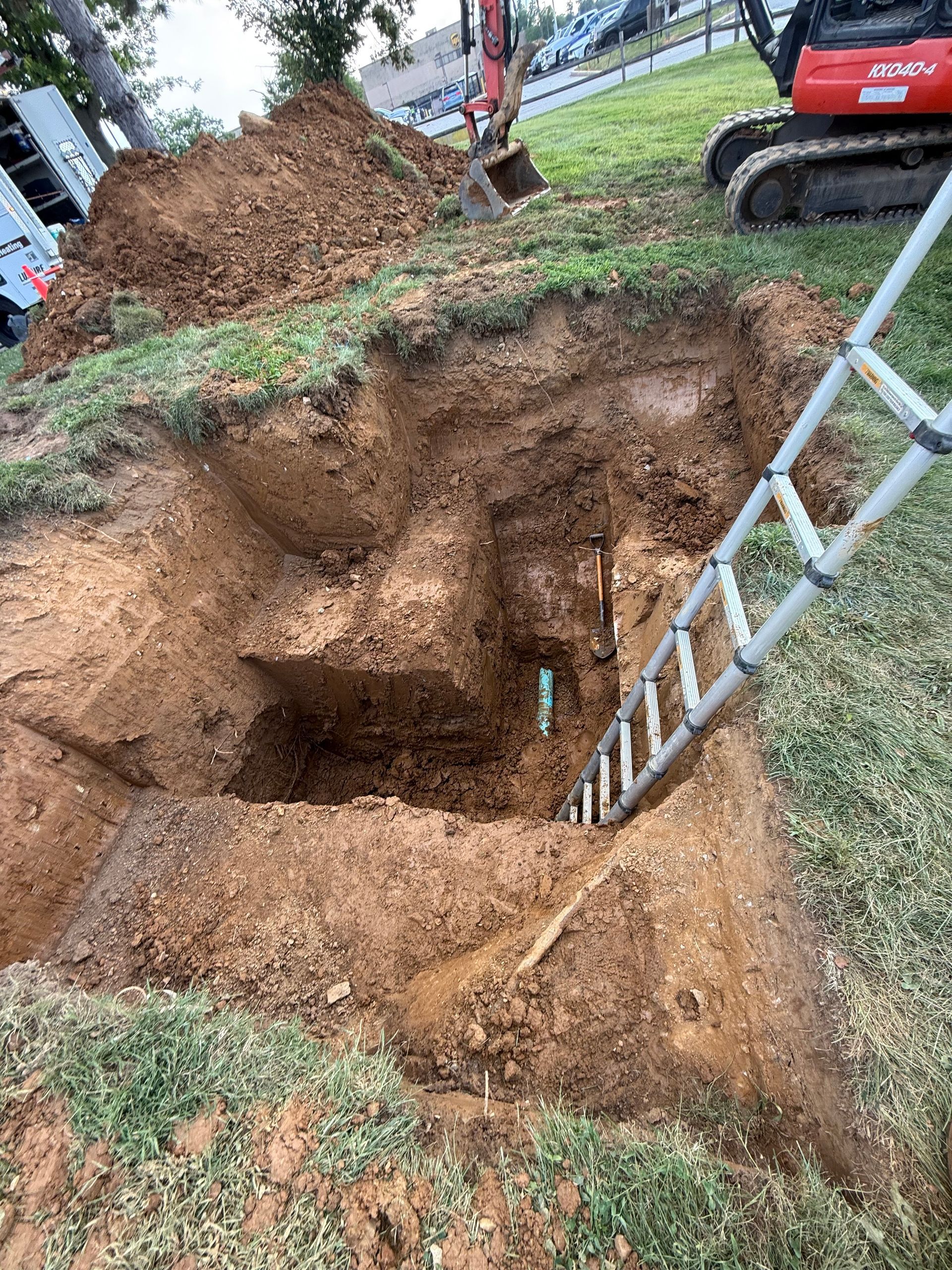 Excavated pit with ladder, red excavator in the background, on a grassy yard.