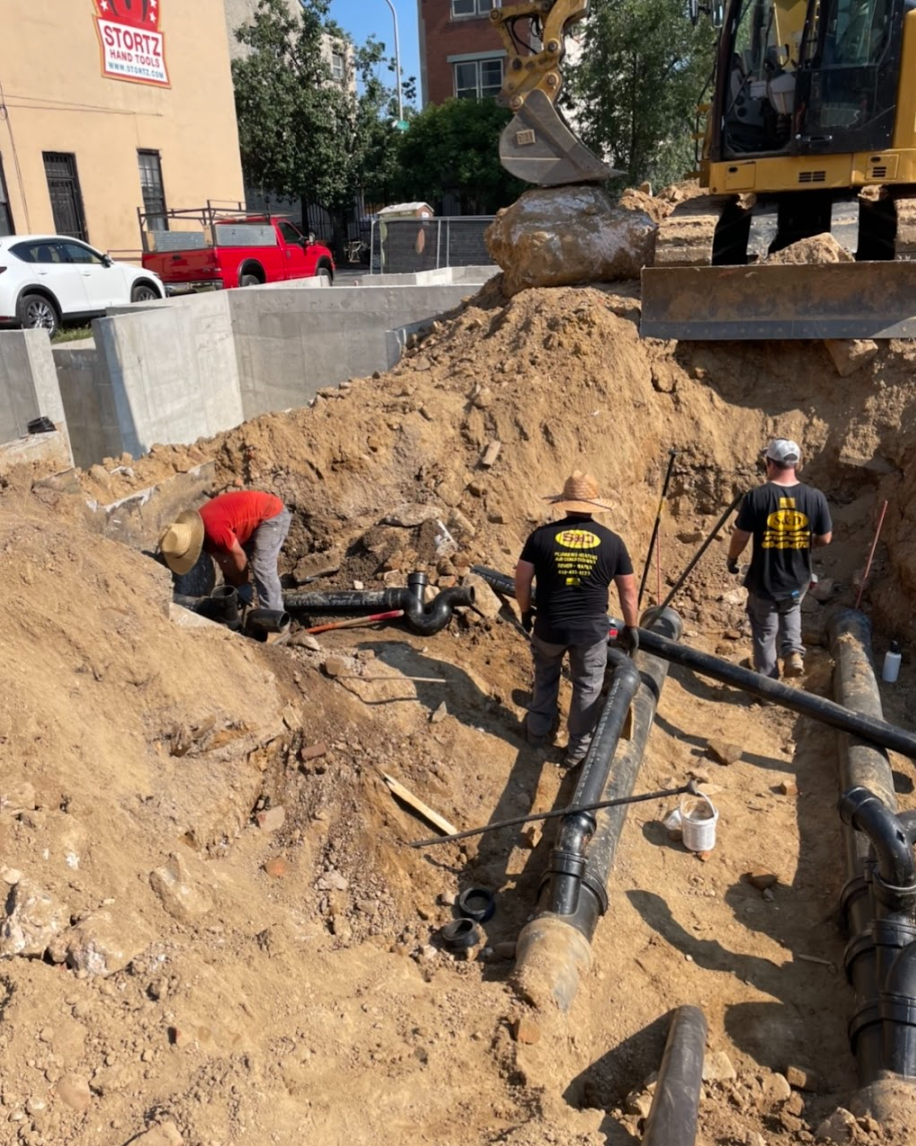 Construction workers in a dirt trench installing pipes with an excavator in the background.