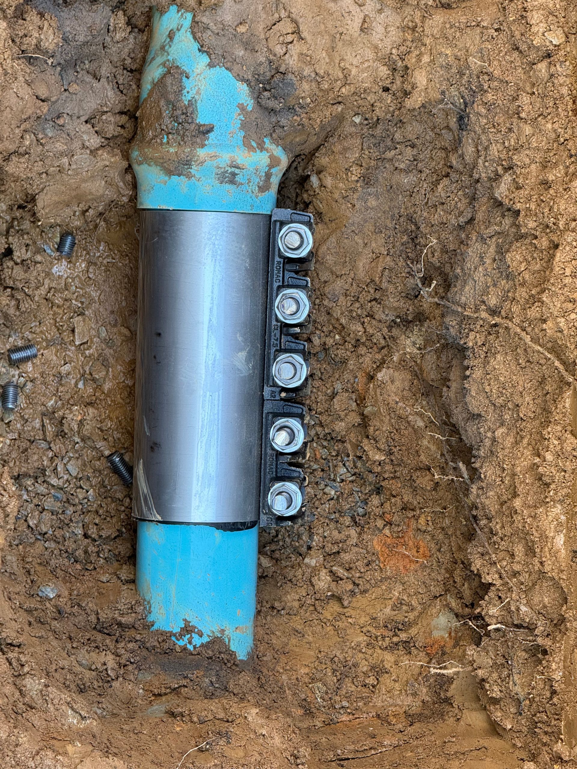 A repaired blue PVC pipe in a dirt trench, held together by a metal clamp with bolts.