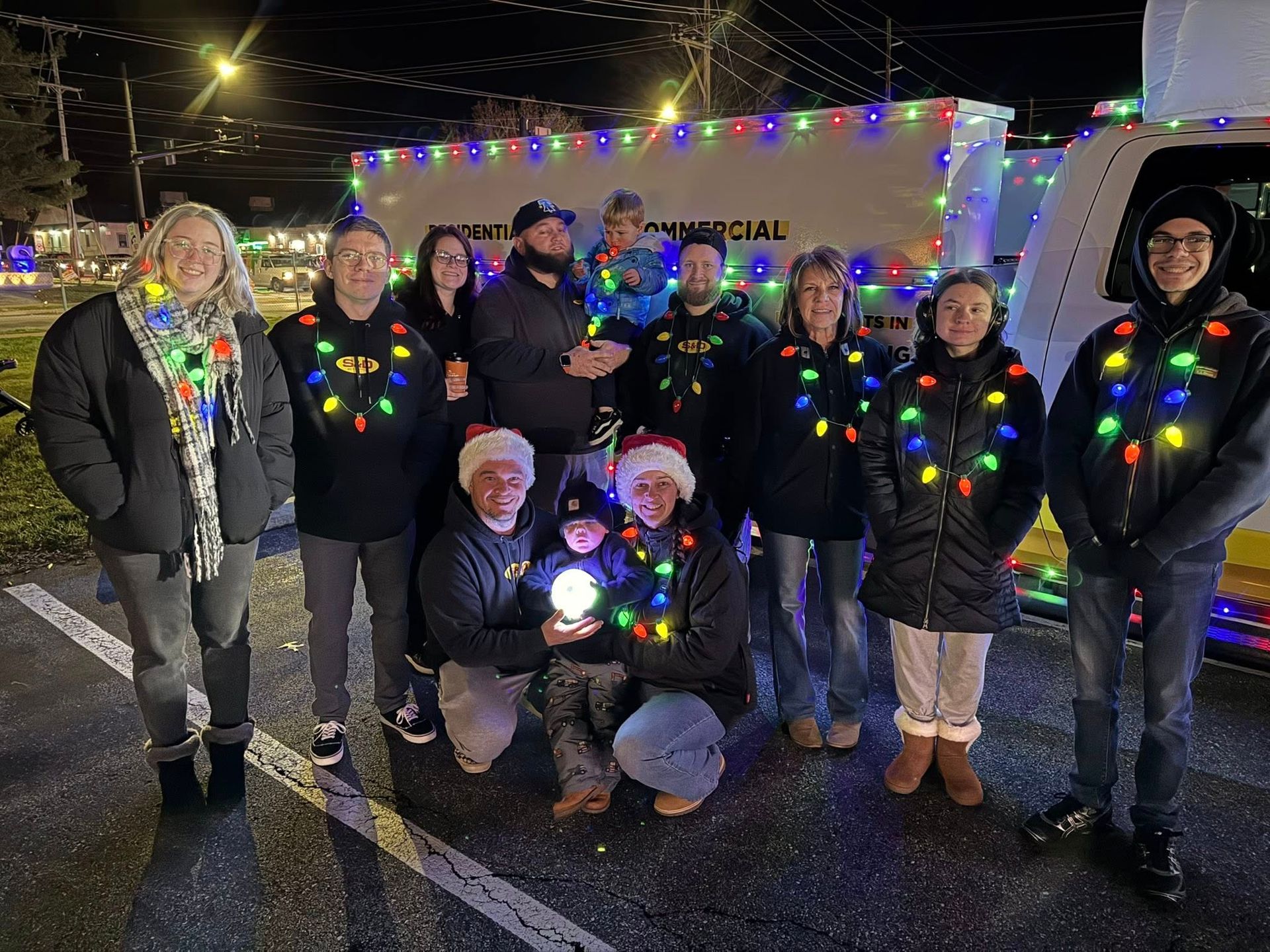Group of people in dark jackets, string lights, and Santa hats pose by a decorated vehicle at night.
