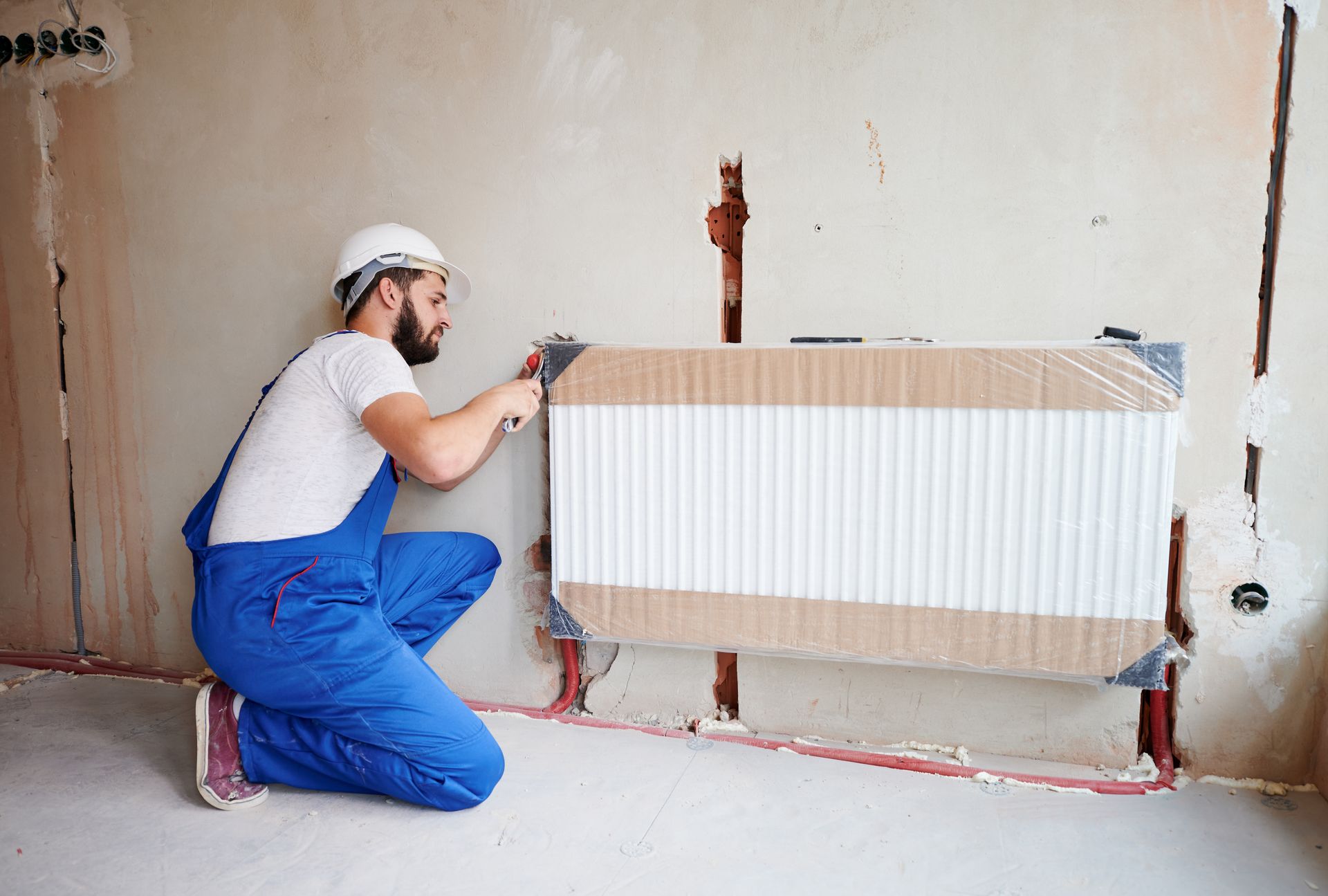 A person in blue overalls installing a radiator on a wall in a room.