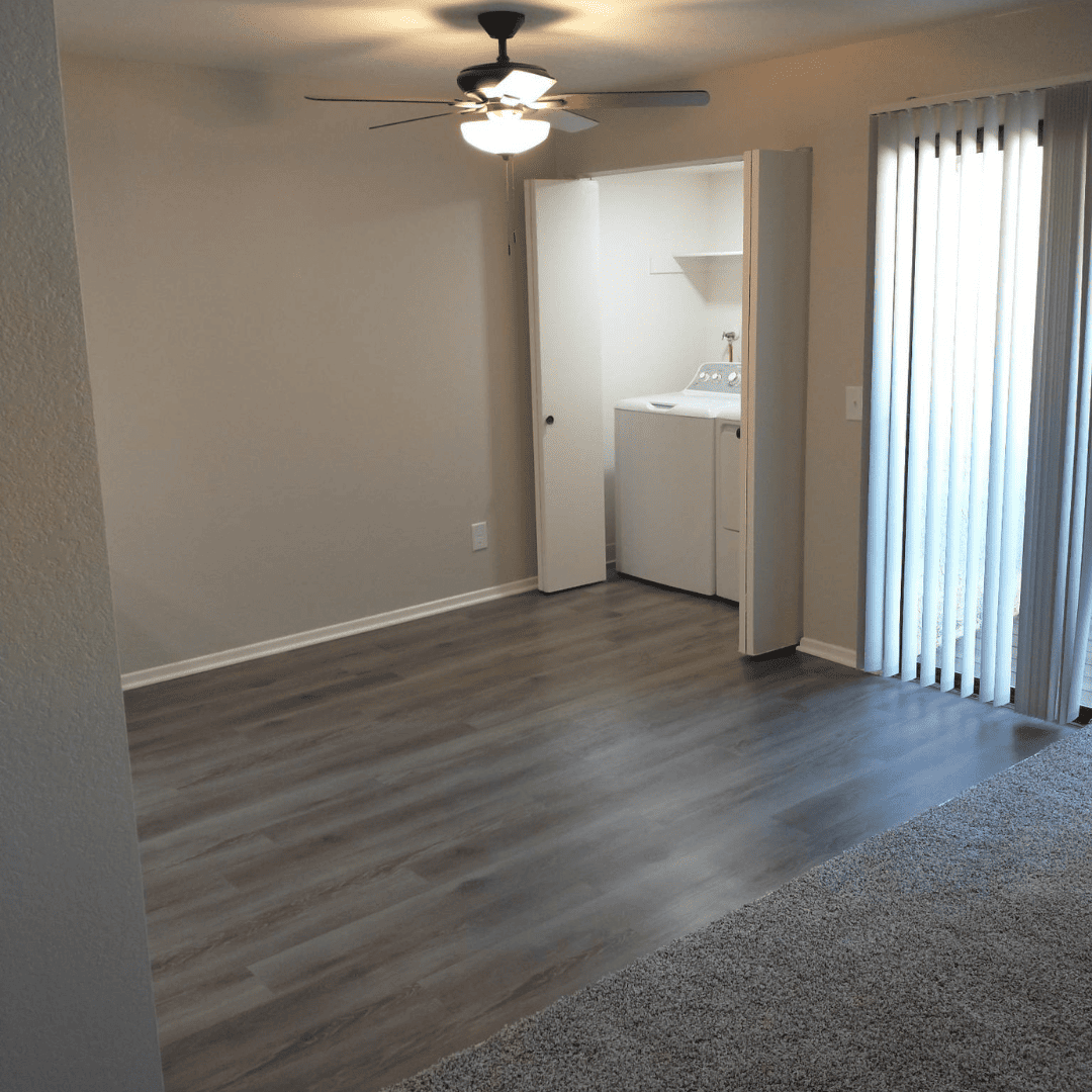 Empty living room with wood-look flooring, laundry closet, and sliding glass door with vertical blinds.