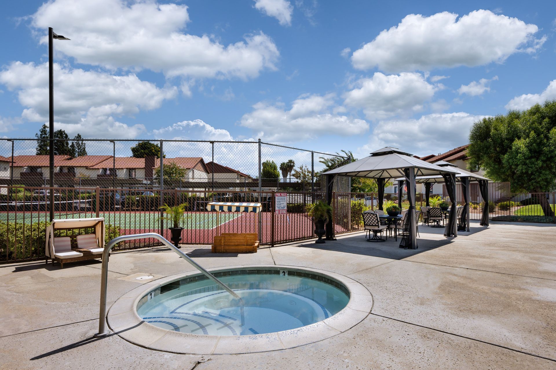 A hot tub in an outdoor area with a gazebo, tennis court, and blue sky with clouds.