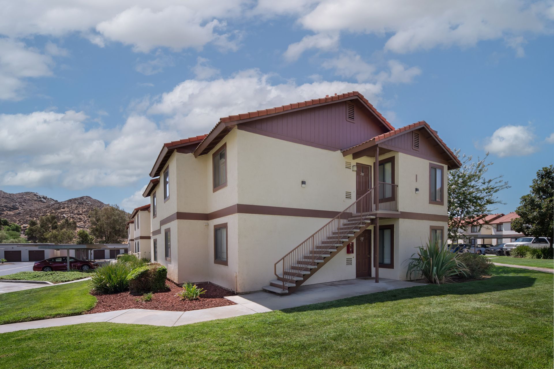 Two-story beige apartment building with brown trim, stairs, and a green lawn under a blue sky.