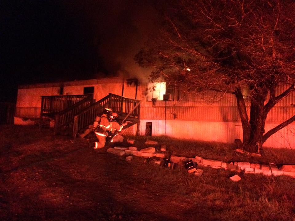 A fire fighter is walking beside a mobile home that is on fire.