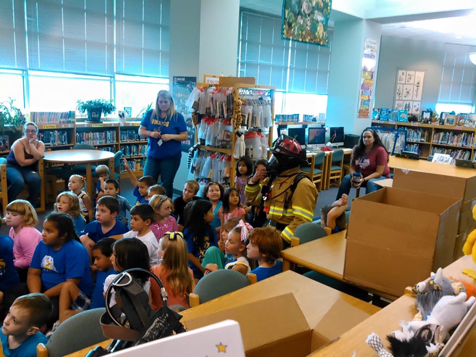 A group of children are sitting at tables in a library watching a fireman