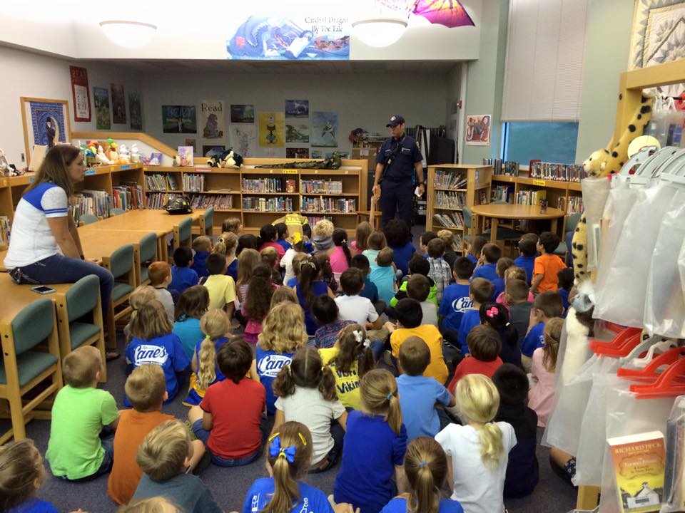 A group of children are sitting on the floor in a library listening to a fire fighter.