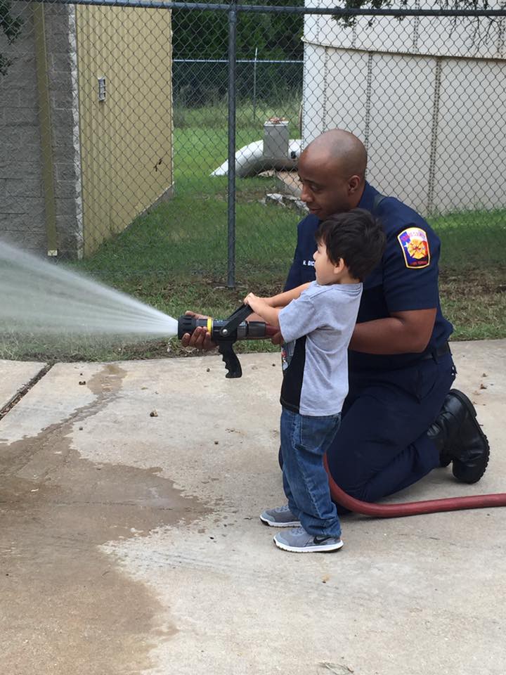A fireman is kneeling down next to a little boy who is holding a fire hose.