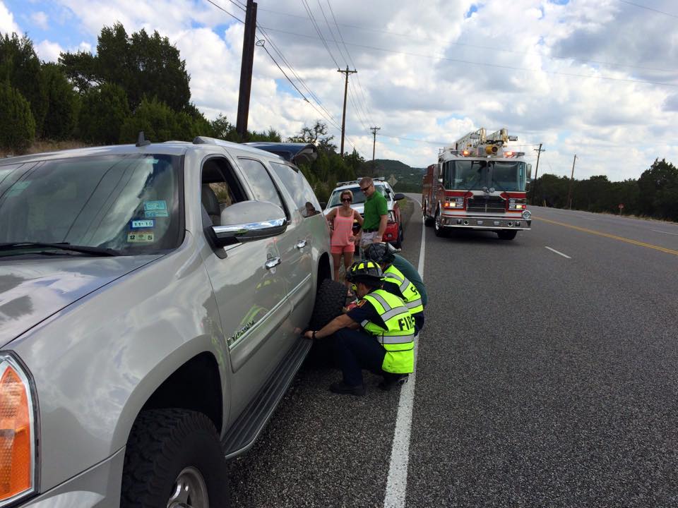 A fire truck is parked on the side of the road while fire fighters help change a flat tire.