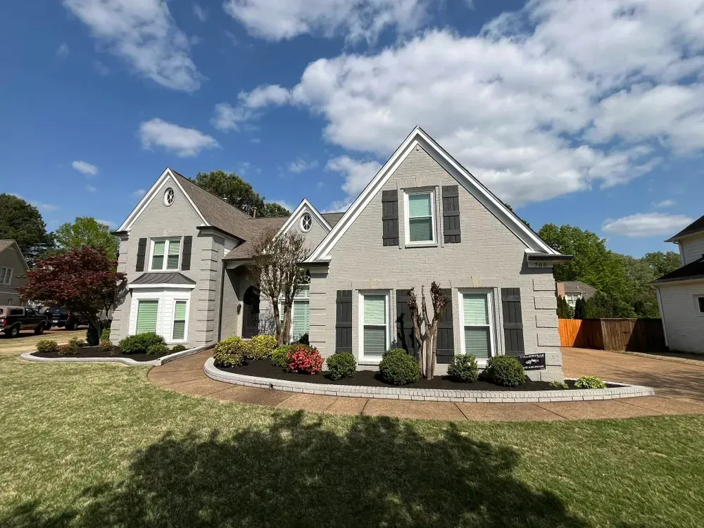 Gray house with black shutters and front yard landscaping under a blue sky.