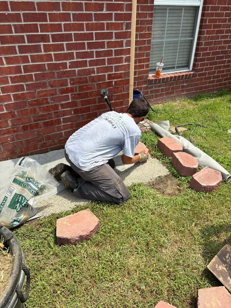 A person kneels laying bricks on a concrete slab near a brick wall, grass, and window.