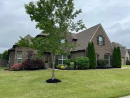 Brick house with brown roof, surrounded by green lawn and landscaping. Cloudy sky.