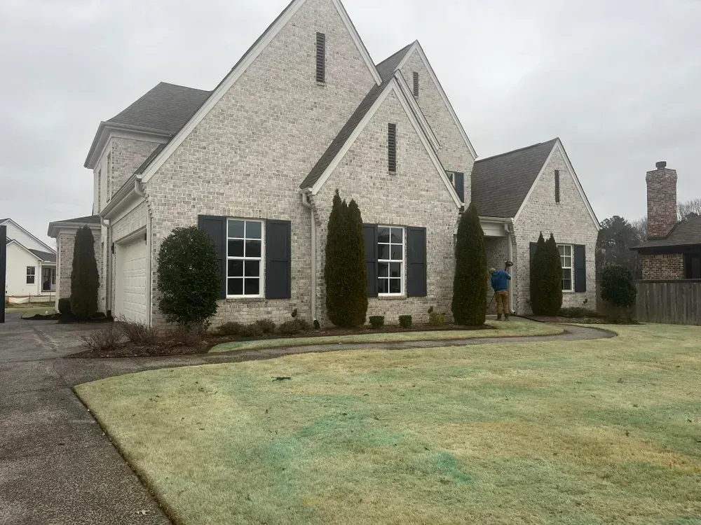 A two-story brick house with dark shutters and a man standing near the front door on a cloudy day.