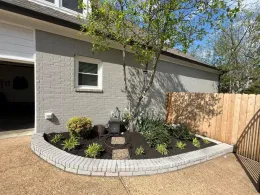 Gray brick-bordered garden bed with black mulch, plants, and a small tree in front of a gray brick house.