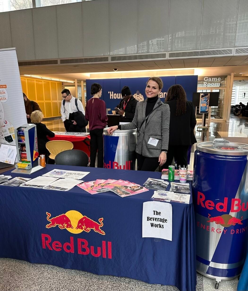 Woman at Red Bull booth, smiling, in a hall. Table with Red Bull branding, cans.