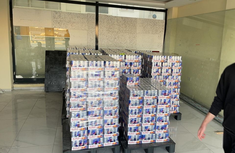 Stacks of canned beverages on pallets inside a building. A person walks by on the right.
