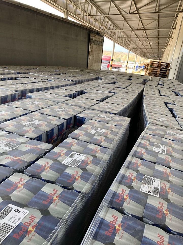 Rows of shrink-wrapped Red Bull cases fill a cargo space, lit by sunlight at the end of the aisle.