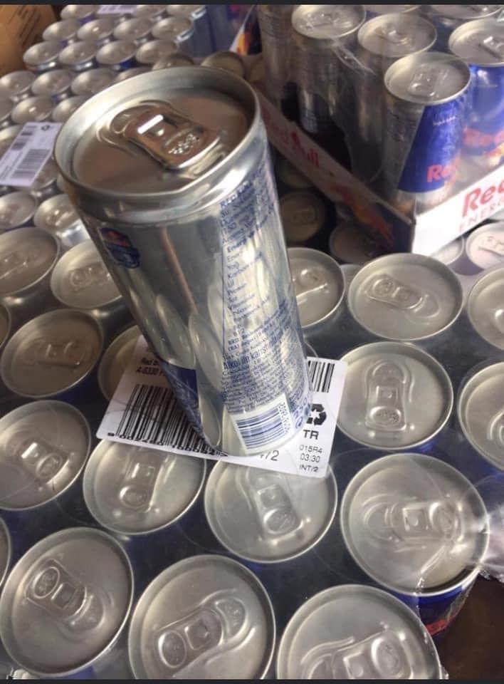 Red Bull cans, some upright, many lying flat. Mostly silver, with blue and red branding, a barcode is visible.