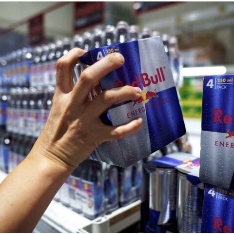 Hand holding a four-pack of Red Bull energy drink in a store, other cans and shelves in the background.