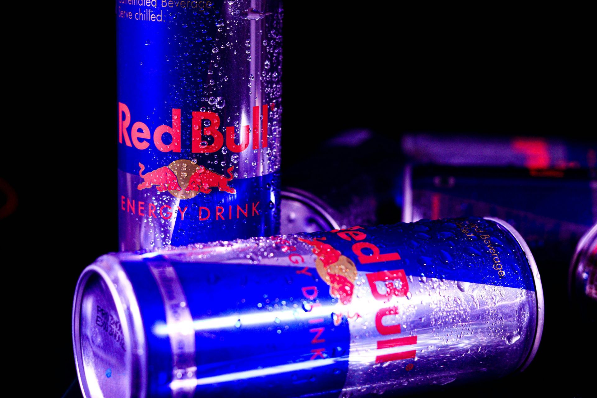 Red Bull energy drink cans, blue and silver, with water droplets on a black surface.