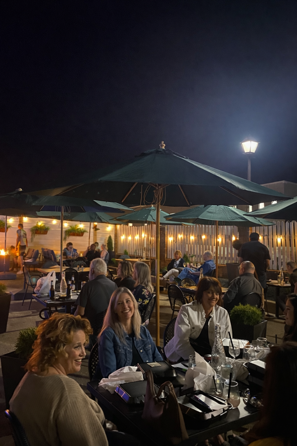 Outdoor restaurant patio at night with diners seated under a large umbrella and warm string lights.