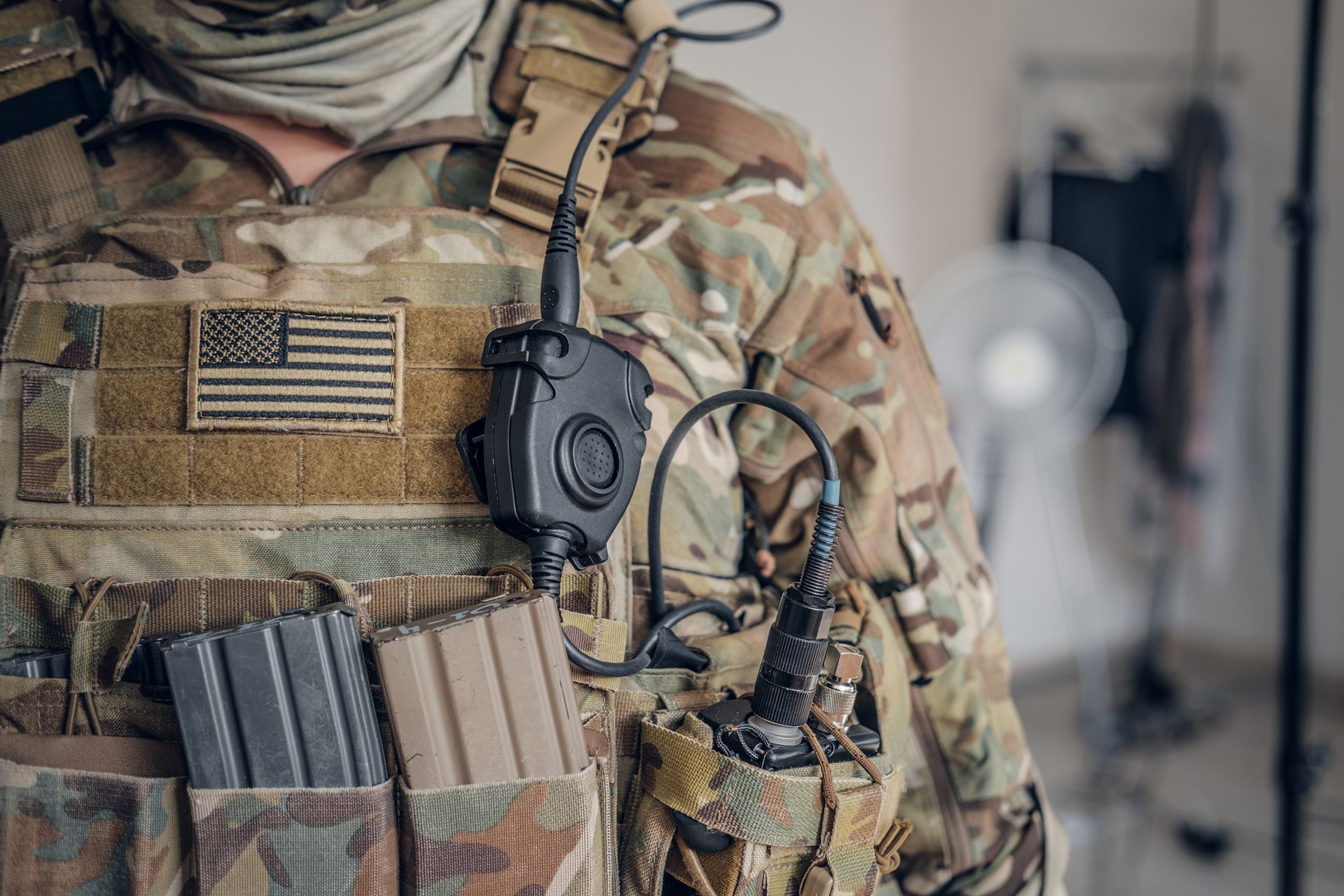 Soldier in camouflage gear with a tactical vest, radio, and American flag patch.