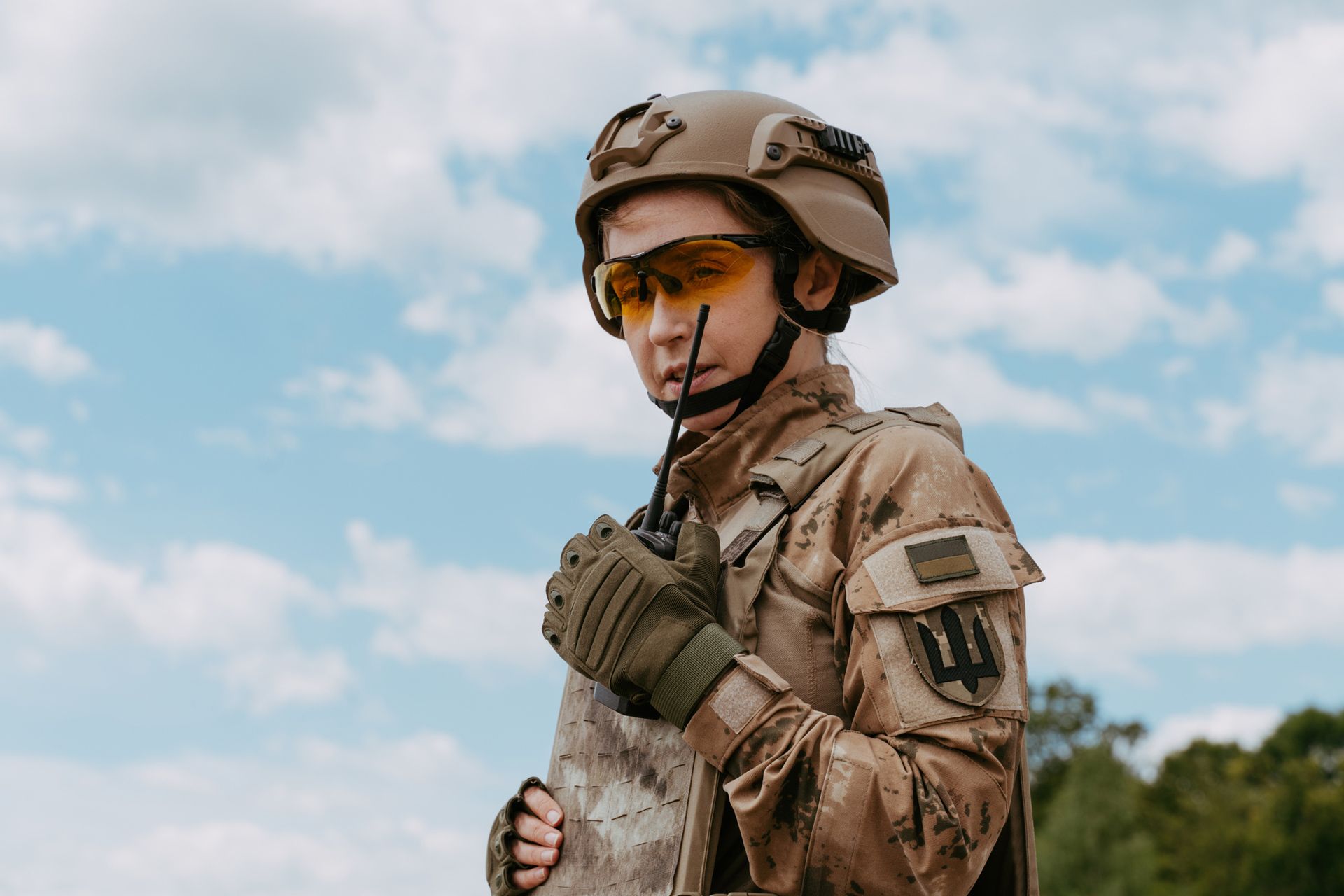 Soldier in tan uniform and helmet holding a radio, with a Ukrainian flag patch, against a blue sky.