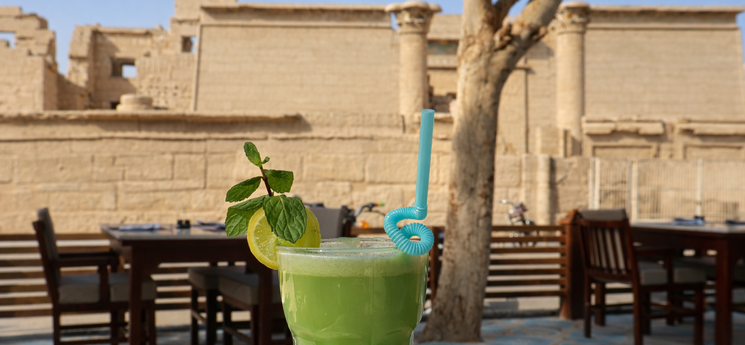 Green drink with lemon and mint, blue straw, in front of a stone building, tree, and wooden tables.
