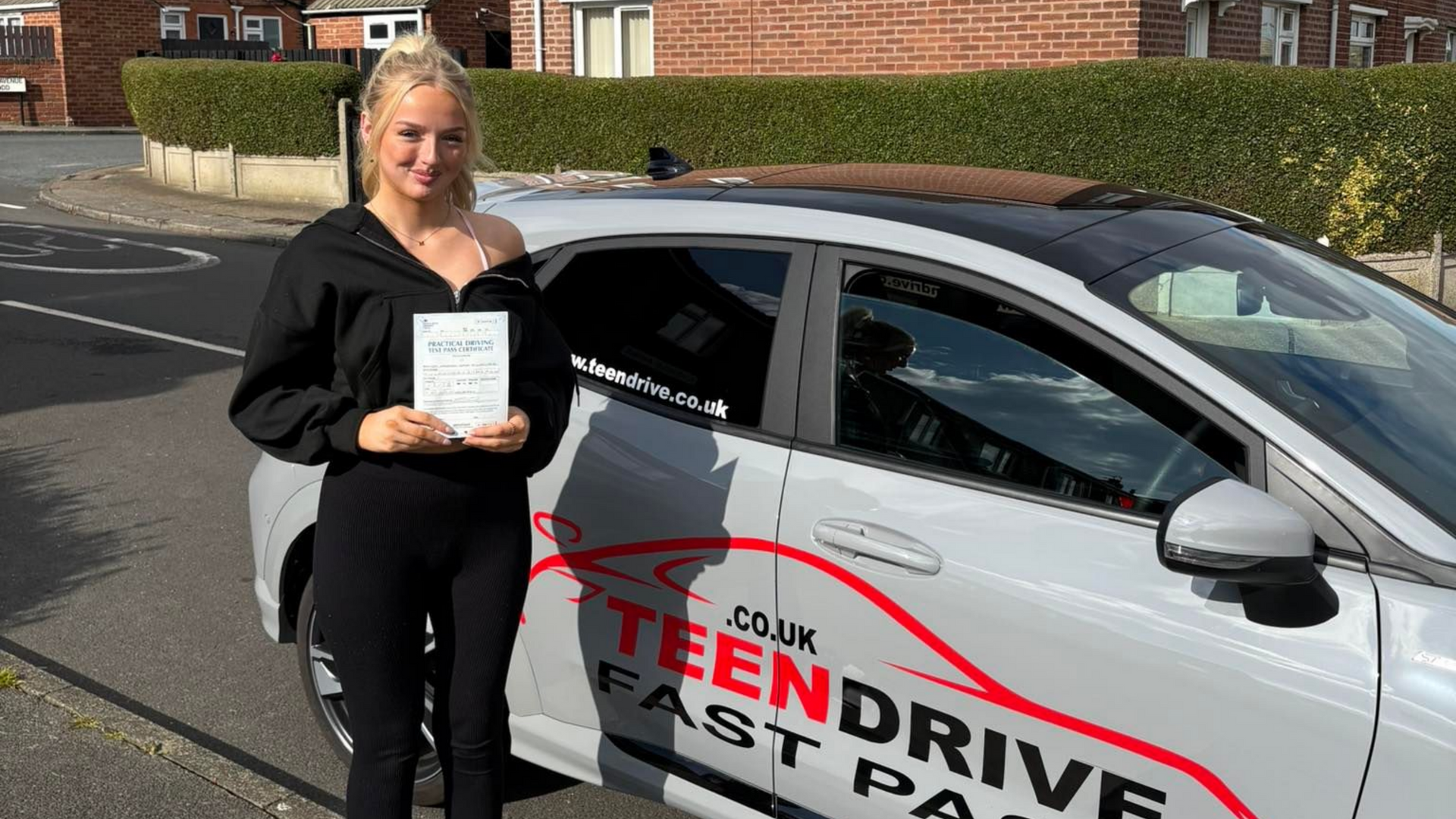 Young woman with driver's license stands by a car with "Teen Drive Fast Pass" on it, in front of houses.