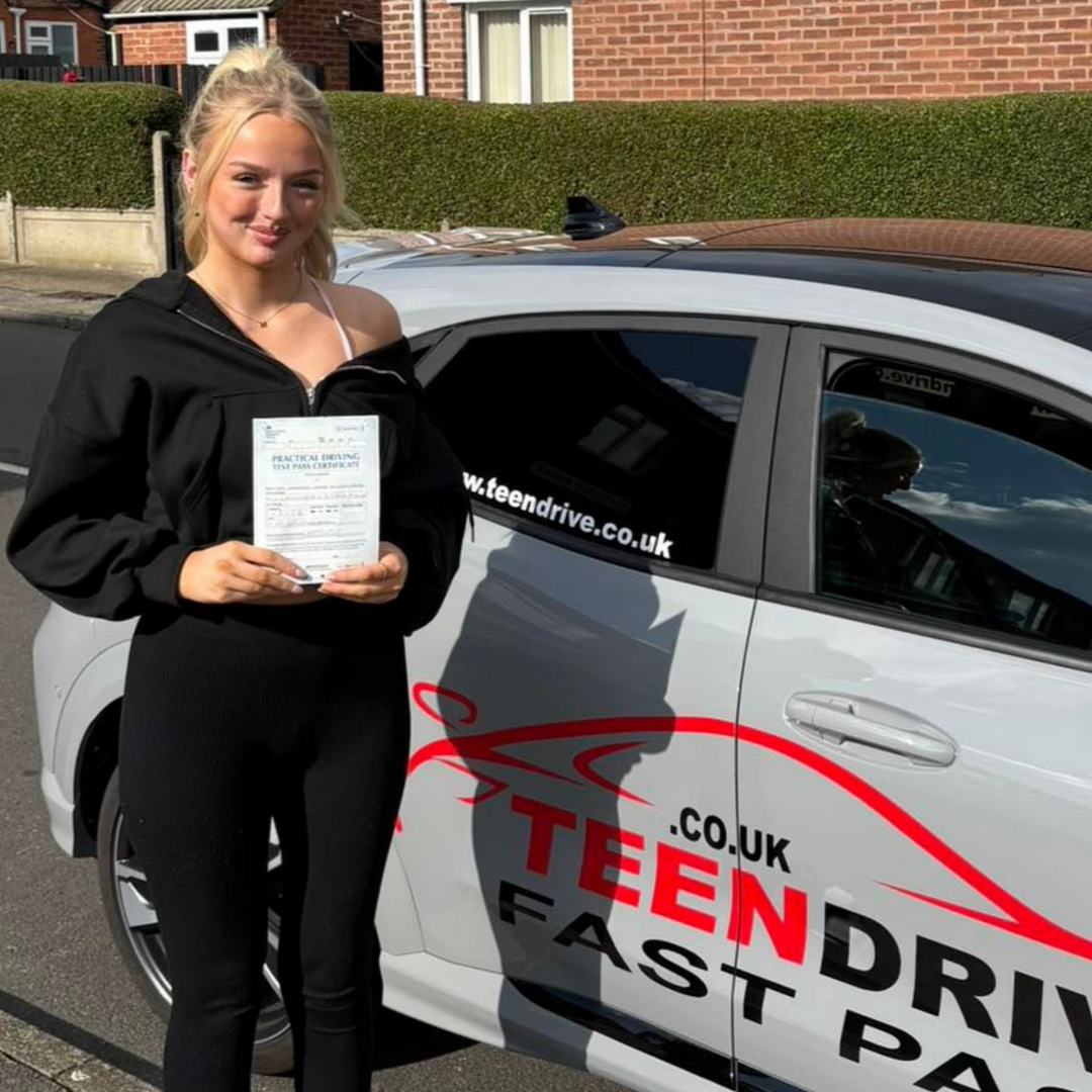 Young woman with driver's license stands by a car with "Teen Drive Fast Pass" on it, in front of houses.
