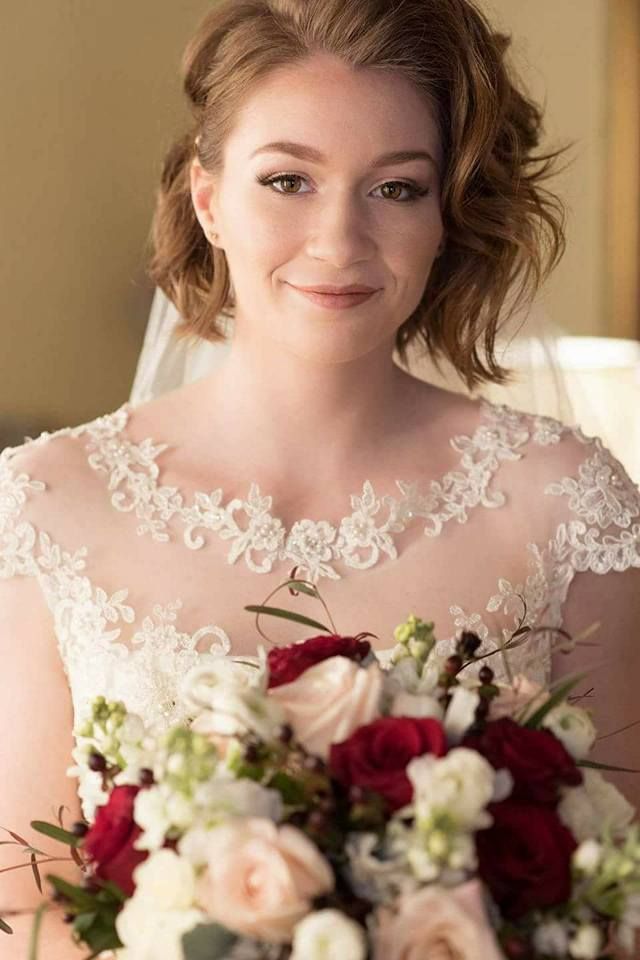 Bride holding bouquet, wearing white lace gown. Smiling, short brown hair.