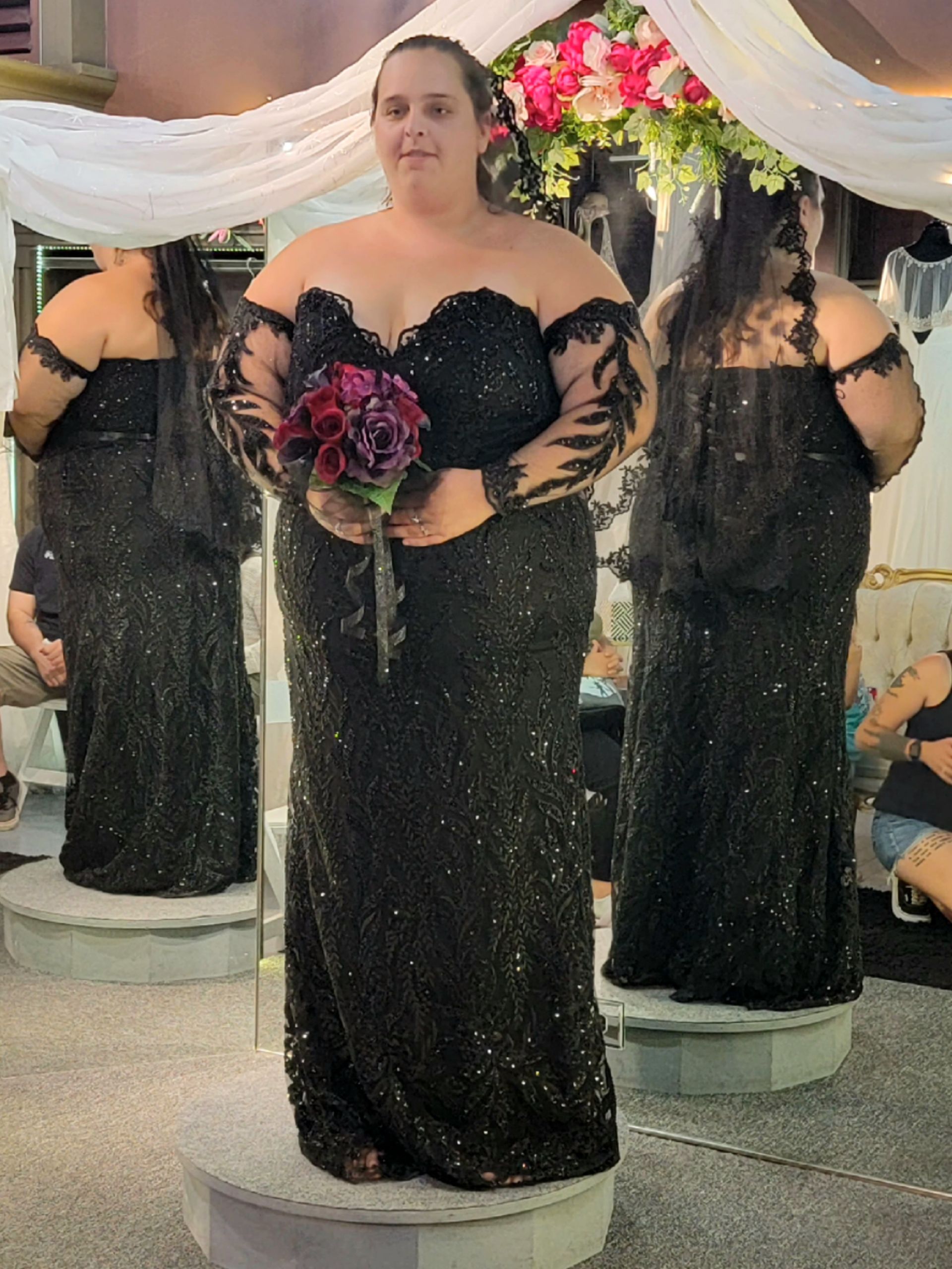 Woman in a black sequin off-the-shoulder wedding dress, holding flowers, stands in front of mirrors.