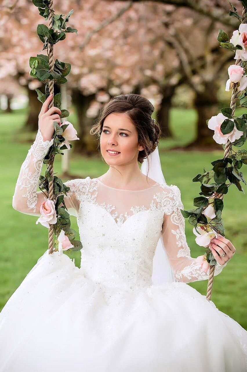 Bride in a white gown sits on a swing decorated with flowers, smiling. Cherry blossoms in the background.