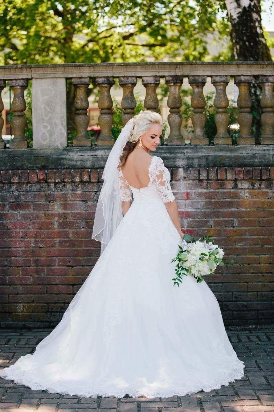 Blonde bride in white dress with veil, holding flowers, standing near a brick wall and balustrade.