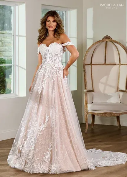 Woman in a white and blush off-the-shoulder wedding dress, posed in a room with a decorative chair.