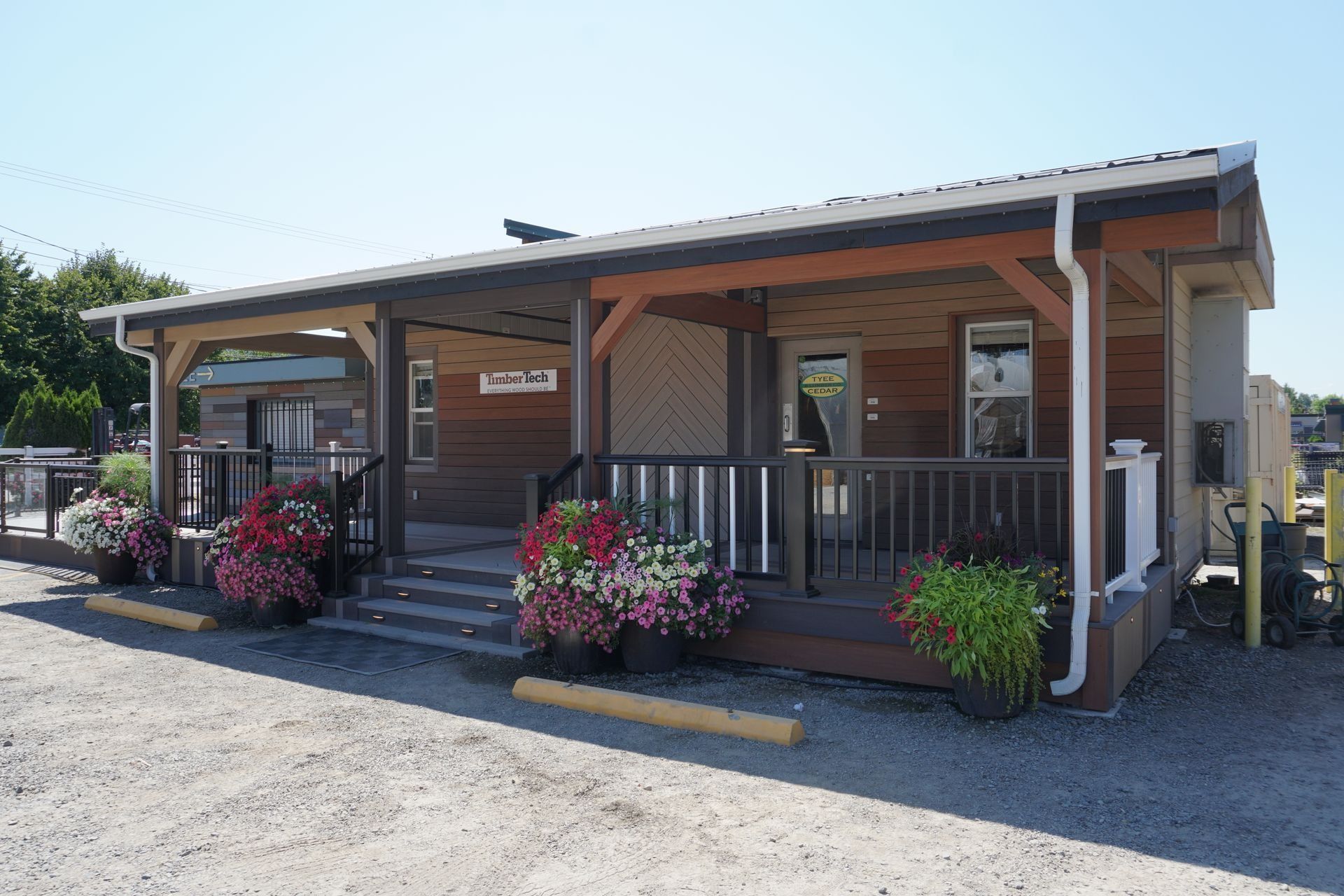 Brown cabin-style building with porch, planters of flowers, and a gravel parking lot on a sunny day.