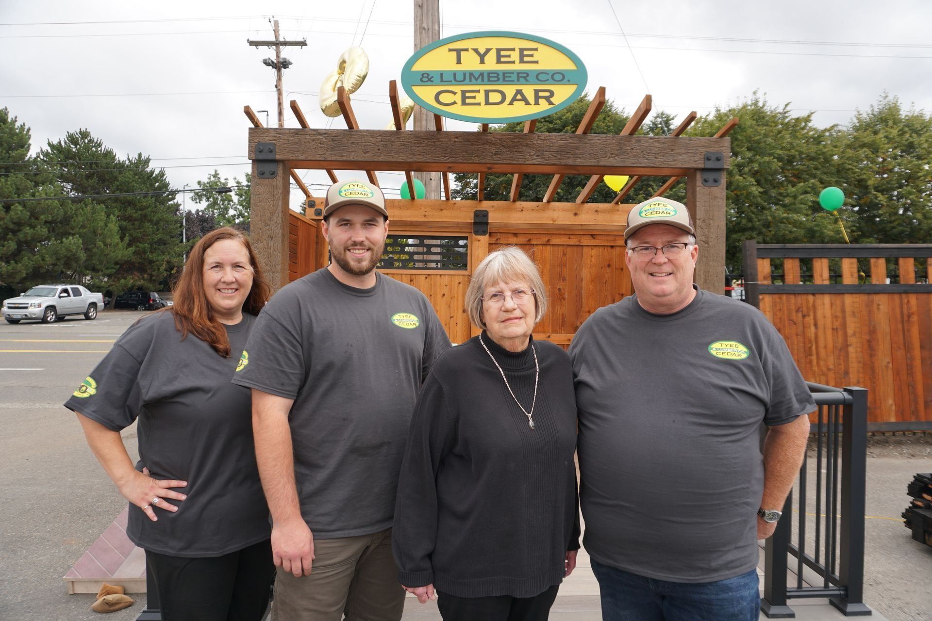 Four people wearing matching shirts and hats pose in front of a lumber store entrance.