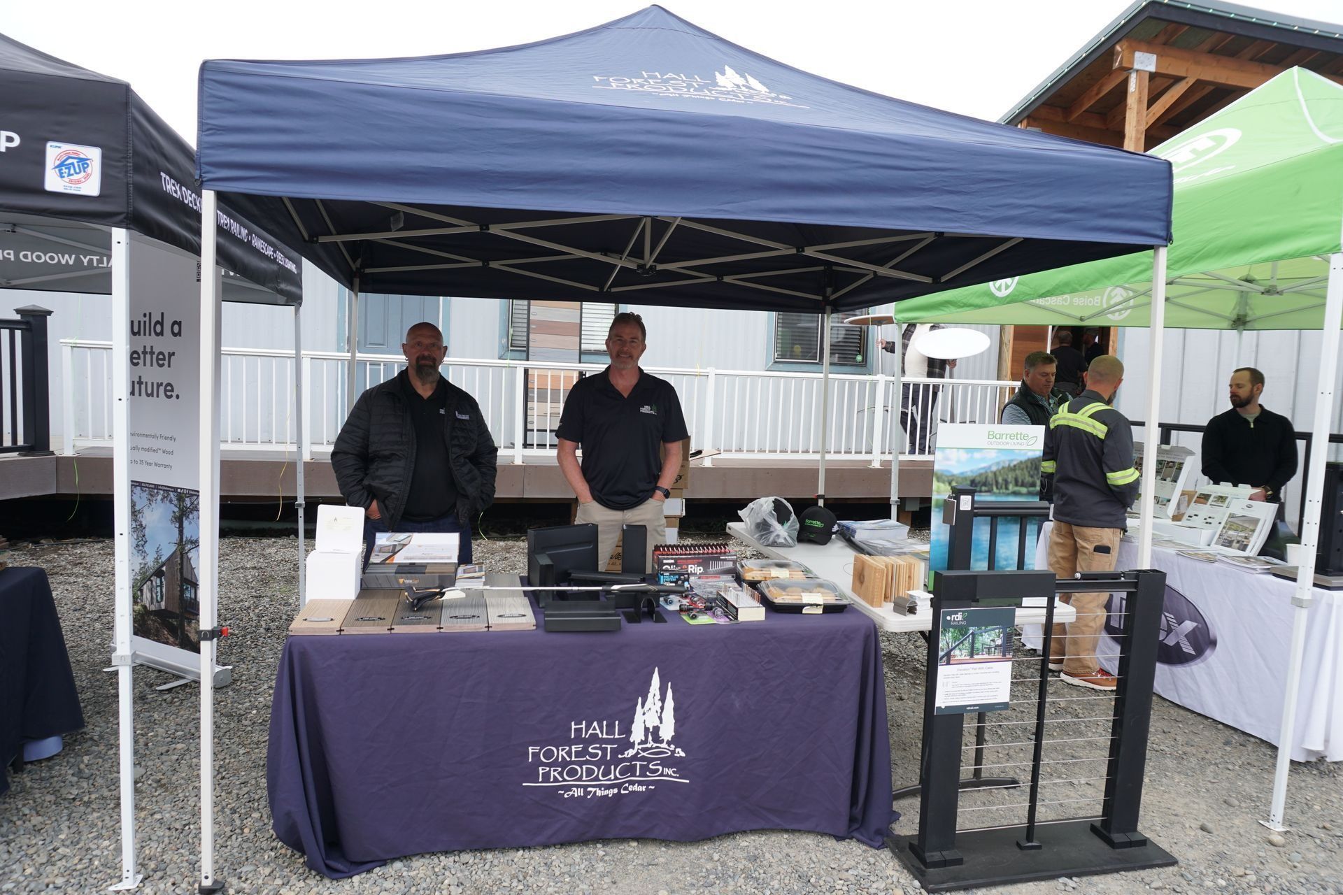 Two men stand behind a booth under a blue tent, displaying samples and brochures at an outdoor event.